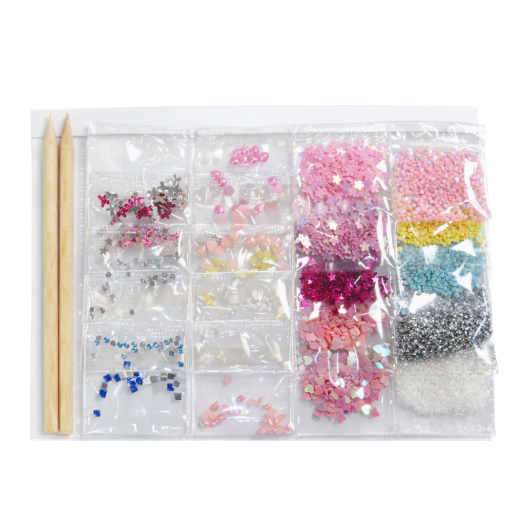 A clear plastic organizer pouch containing multiple compartments filled with assorted nail art embellishments, rhinestones, glitter, beads, and small decorative pieces in pink, silver, yellow, blue, white, and clear. Two wooden sticks are placed beside the pouch on a white background.