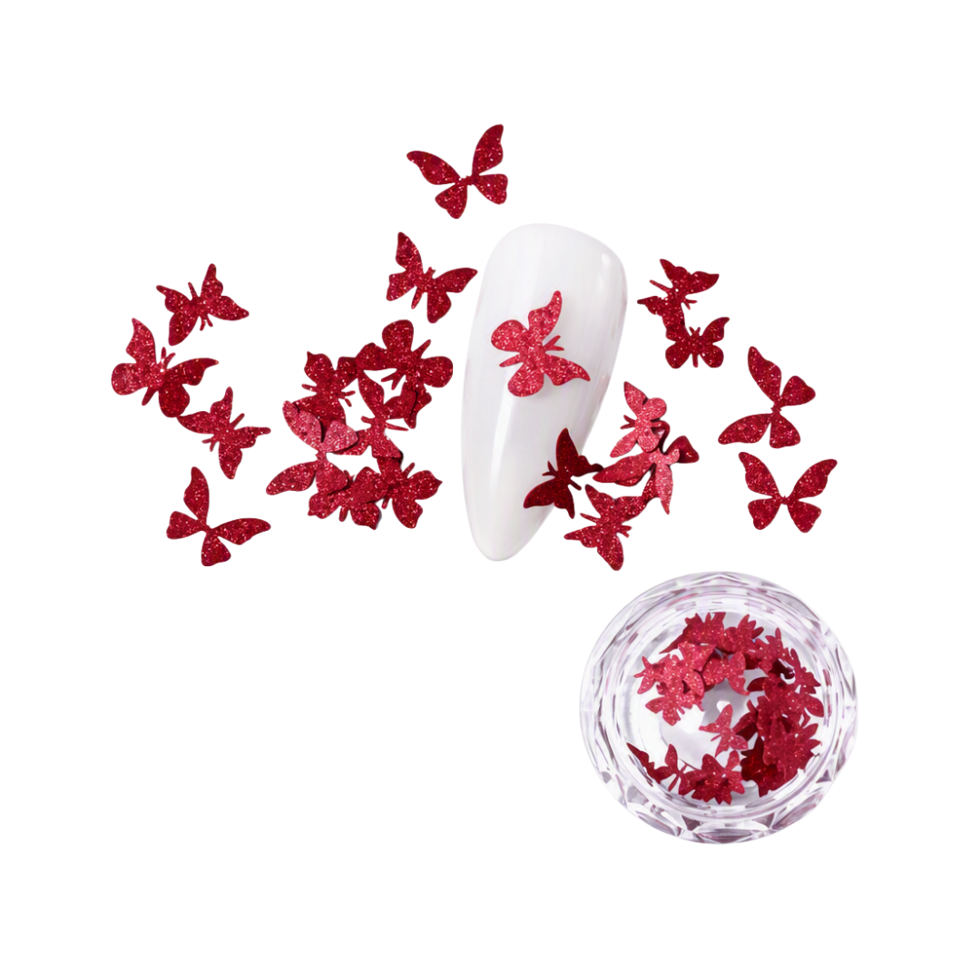 Red glitter butterfly nail art decorations scattered around a white nail tip, with a small clear round dish of additional red butterfly flakes on a white background.