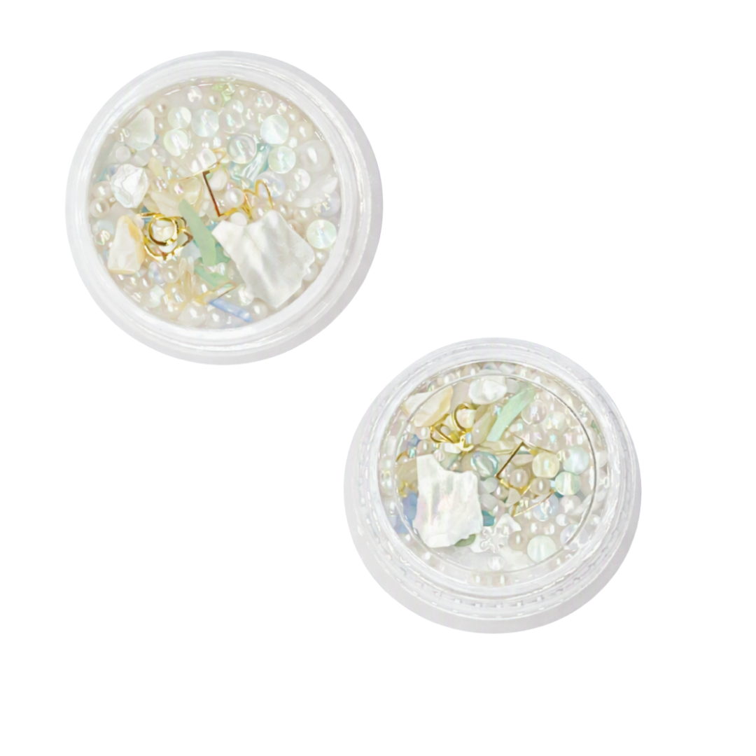 Two small round clear plastic jars filled with decorative nail art embellishments, including iridescent pearls, white flakes, gold foil, and pale green accents, shown on a plain white background.