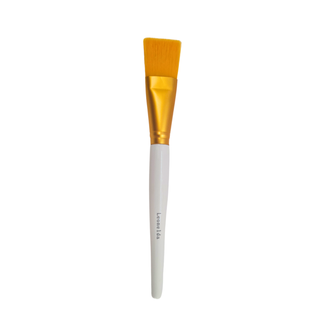 A flat facial mask brush with orange-yellow bristles, a shiny gold ferrule, and a white handle with the brand name 'Leonelda' printed on it, set against a plain white background.