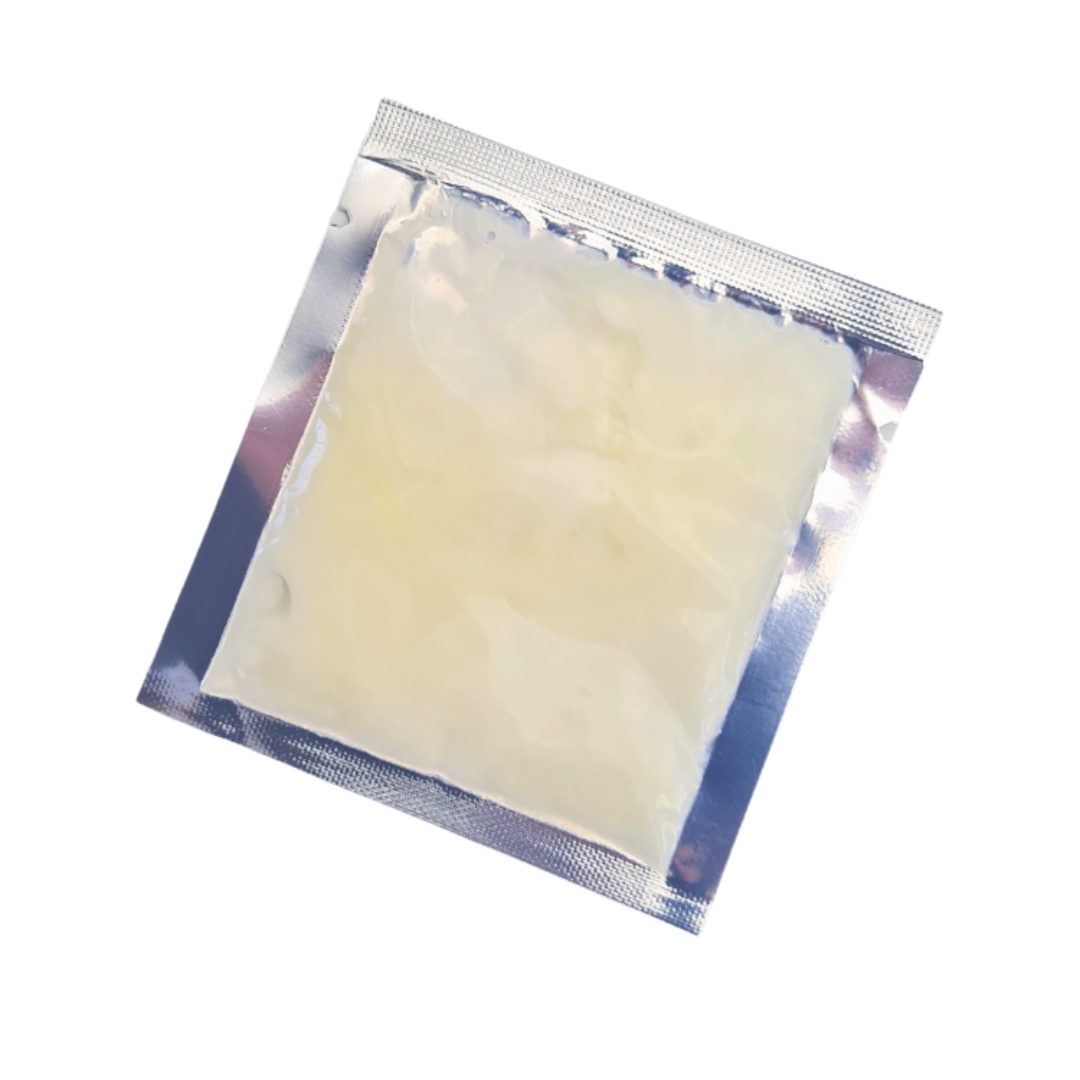 A square translucent off-white pregnancy safe massage balm sealed inside a metallic silver foil packet, shown on a plain white background.