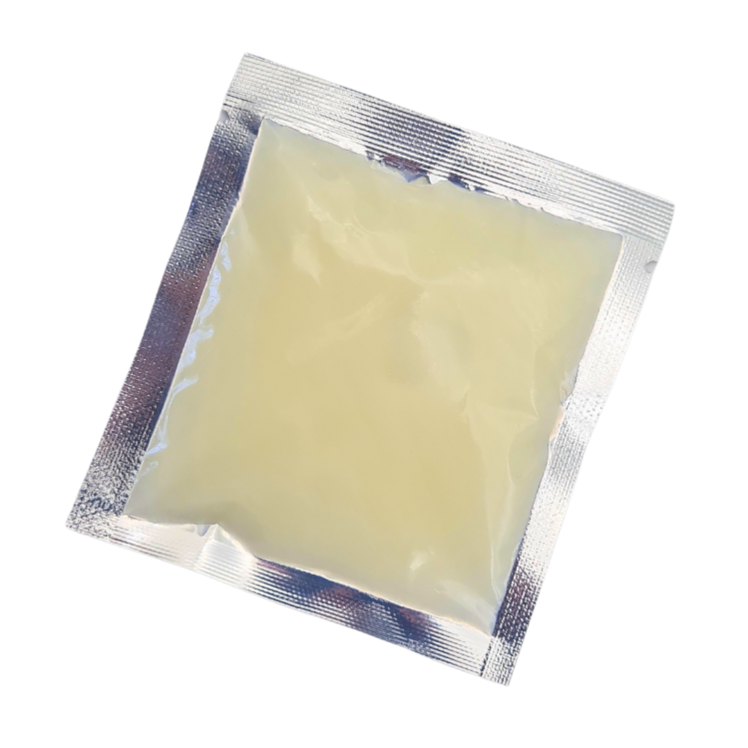 A square silver foil packet containing pale yellow translucent muscle ease full body massage balm, photographed on a plain white background.
