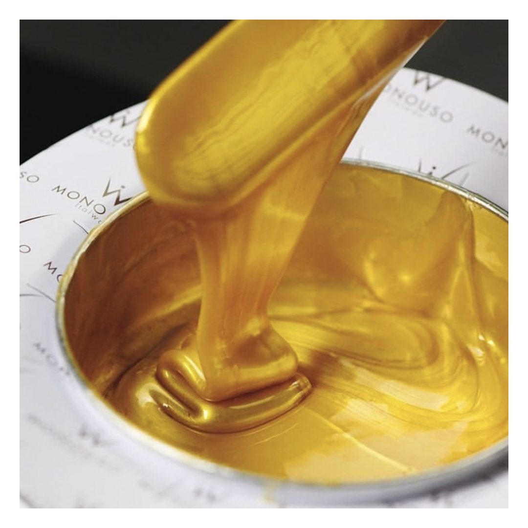 A close-up of thick, glossy metallic gold Italwax film wax being scooped with a spatula from a white tin can, with the brand name partially visible on the tin.