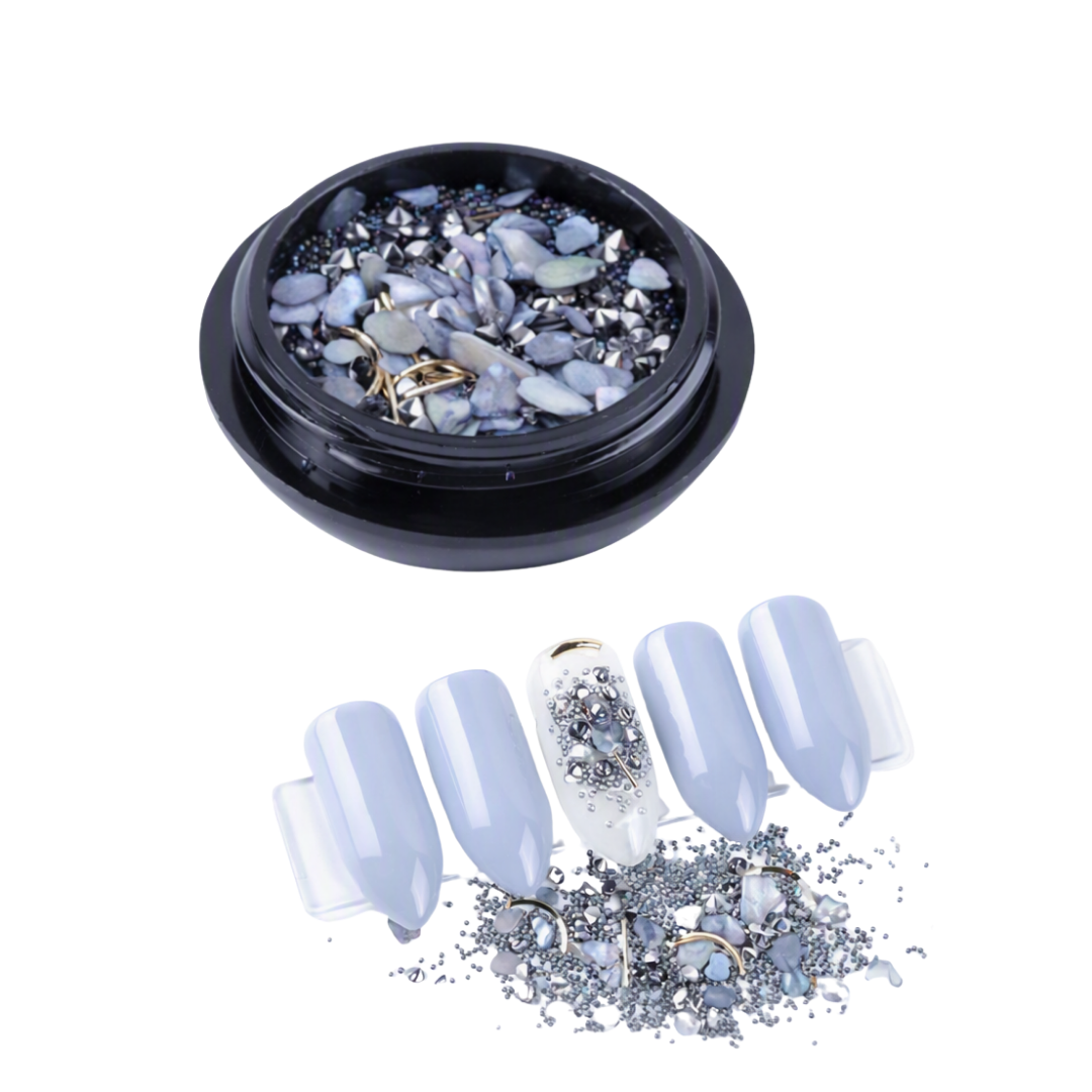 Nail art glitter and stone mix in a round black container, shown above false nails decorated with the same light blue-gray glitter and small gold ring accents on a white background.